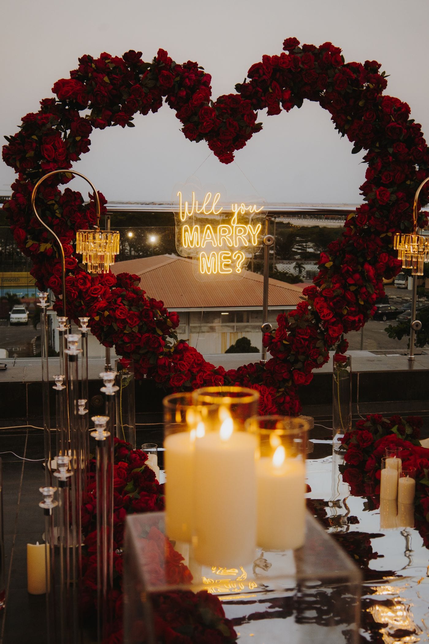 Beachside marriage proposal at sunset with a red-rose heart arch and 'will you marry me' sign